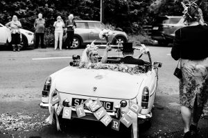 bride and groom drive away in vintage MG while bride celebrates with flowers in the air