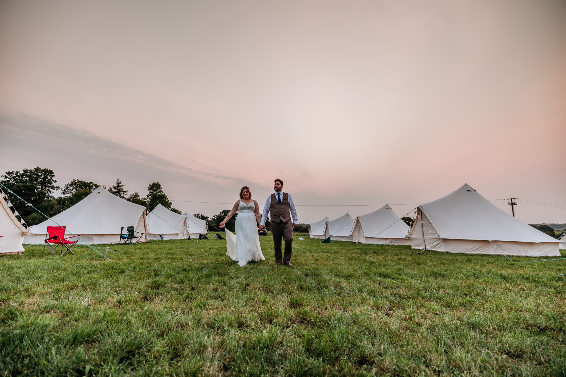 a soft sunset and just married couple walking through the glamping field of bell tents at ashridge great barn