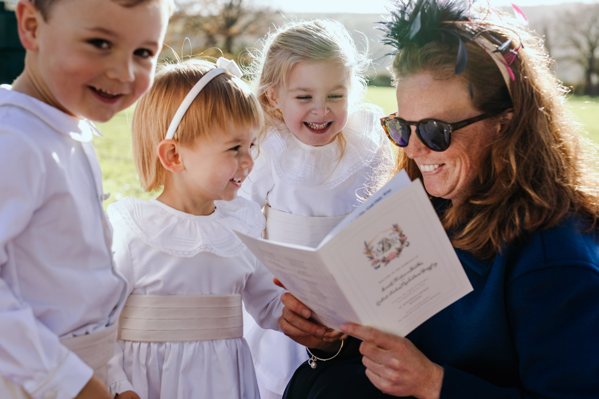 flower girls and paige boys at a bovey castle wedding are entertained by an aunt with a book outside the church in manaton
